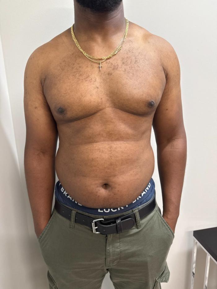 A shirtless man wearing a gold chain, green pants, and a black belt stands with his hands in his pockets against a plain background.