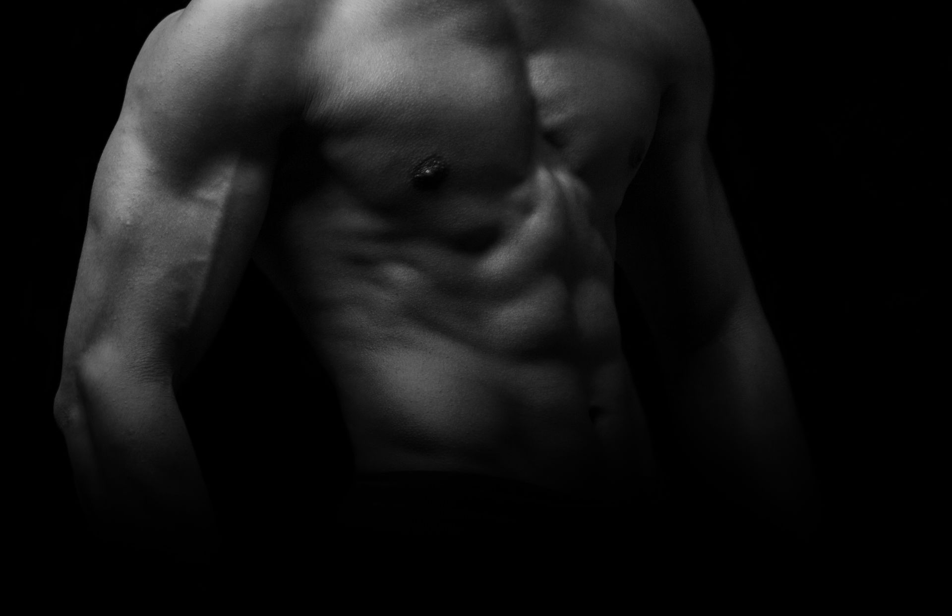 A black and white photo showing a muscular, shirtless male torso and arm, highlighting defined abs and chest against a dark background.