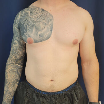 A shirtless man with a detailed black and gray tattoo covering his left arm and upper chest stands against a dark background.