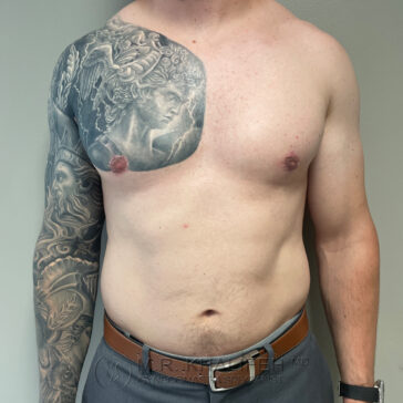 A shirtless man with a large black and grey tattoo covering his left shoulder, upper chest, and arm stands against a plain wall.