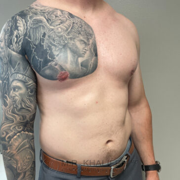 A shirtless man with a large, detailed black and grey tattoo covering his left arm, shoulder, and upper chest, standing against a plain wall.