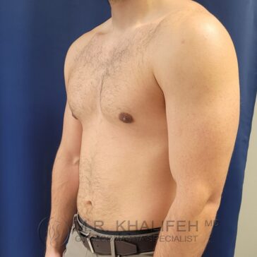 A shirtless man standing in front of a blue curtain, shown from the waist up and turned slightly to the side.
