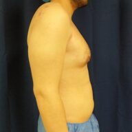 Side profile of a shirtless person standing against a dark curtain, wearing blue pants and a cap.