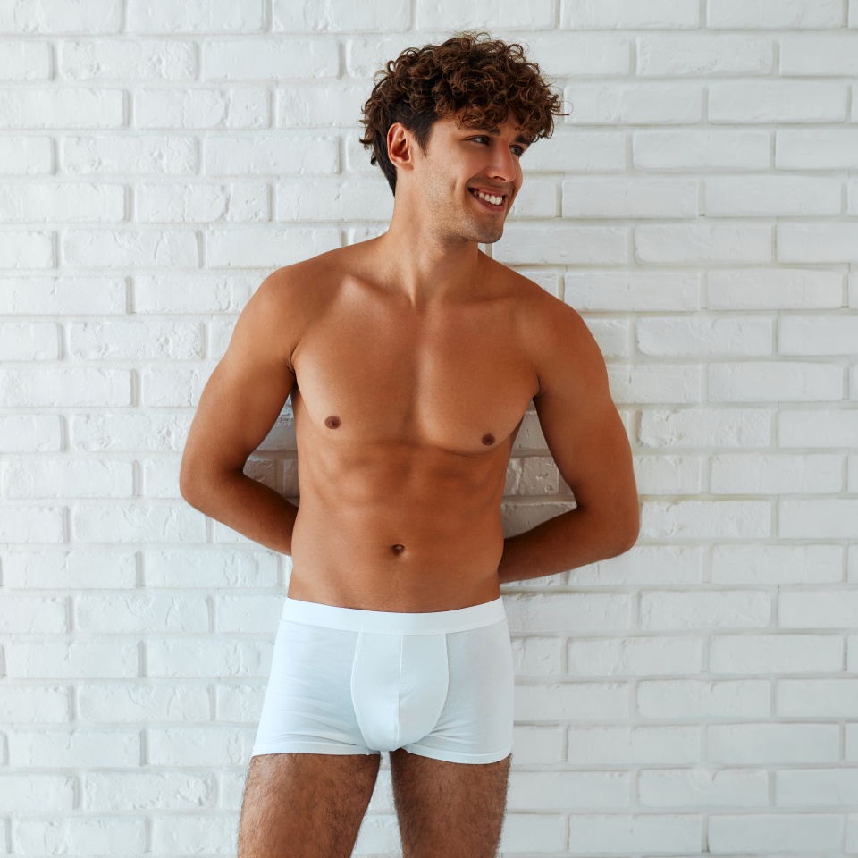 A handsome, muscular, sexy young man in white shorts with a naked torso standing against a white brick wall.
