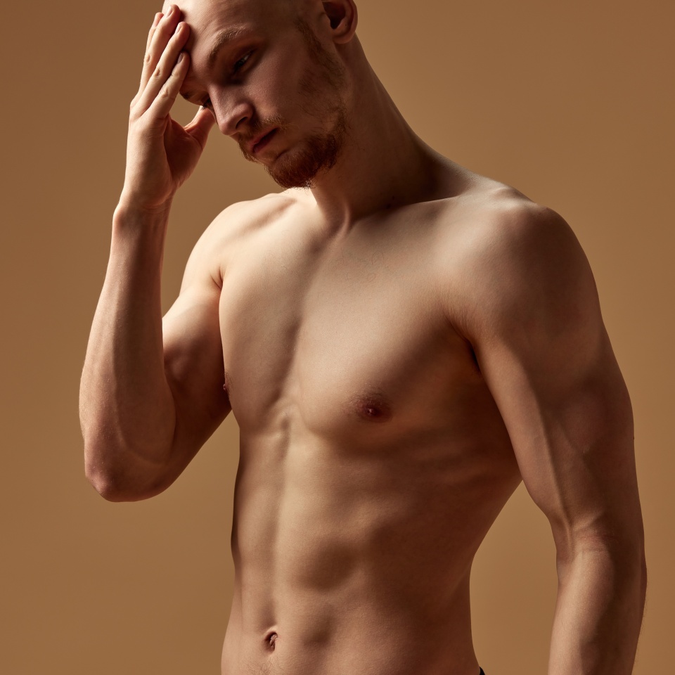 Portrait of pensive bald man posing in underwear holding hand on forehead against beige studio background. Concept of men's health, self care, fashion and beauty, healthy lifestyle. Ad