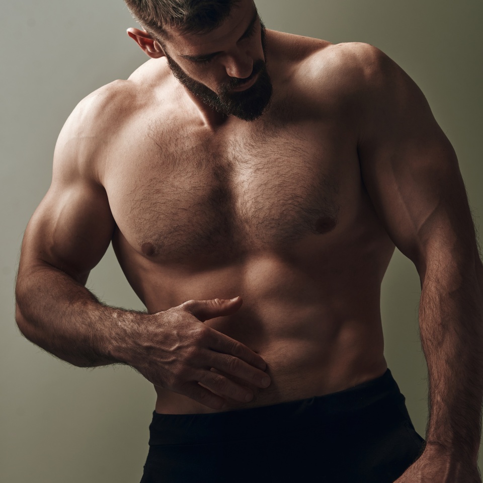 A muscular bodybuilder stands in a dimly lit studio, flexing and emphasizing his toned physique. The dramatic lighting highlights his well-defined muscles, showcasing fitness.