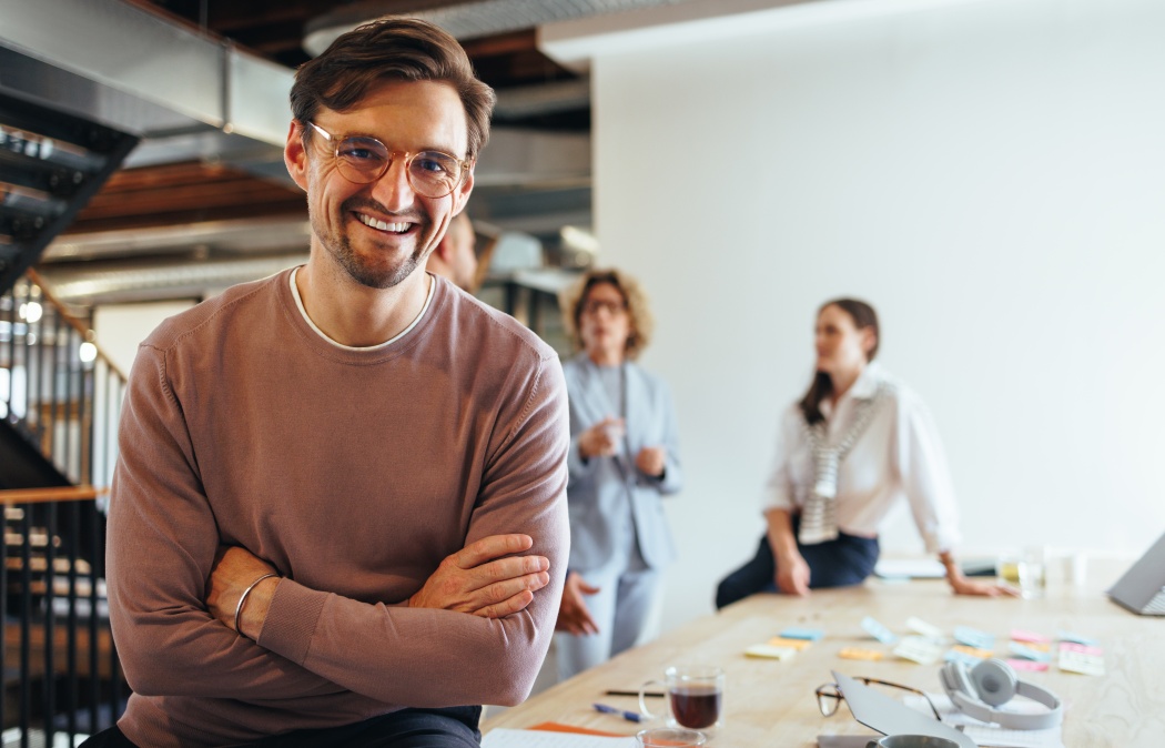 Successful business man sitting in a boardroom with his team in the background. Happy business professional looking at the camera in a meeting. Business people working on a project.